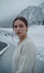 a woman wearing long hair looking off to the side while standing in front of a snow covered mountain
