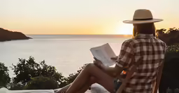 a woman reading a book on a sunny day with the sun peeking over water