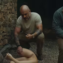 the body of a naked man is being tended by a tattooed man