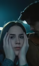 a couple sit together in a car, one looking at the camera and the other a sad