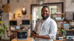 man in white shirt holding up a cell phone,the man is holding a cell phone in a room with many items