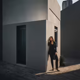 a woman in black leaning against a grey building with her arm on the wall and she is talking on her cell phone