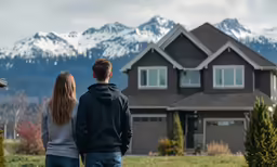 a couple stands in front of their house with snow capped mountains behind them