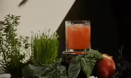 a glass with juice, basil and tomatoes beside a plant