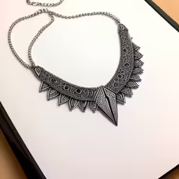 a silver necklace with a decorative design sitting on a paper,this is an elaborate black necklace on a white surface