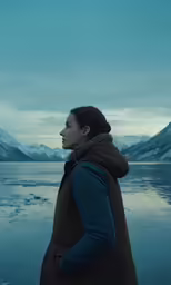 a woman stands with her hands in her pockets at dusk overlooking water