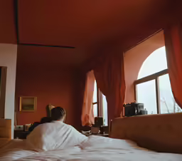a lady sitting in bed next to two large windows