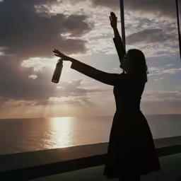 a woman in black is stretching her arms with one hand and holding a bottle of alcohol near a sea