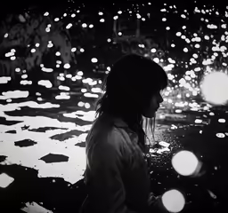 woman standing in a dark puddle, illuminated by lights in the background