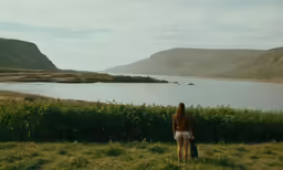 two women stand facing a body of water with mountains in the background