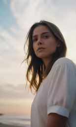 a woman with brown hair and a white shirt standing by the ocean