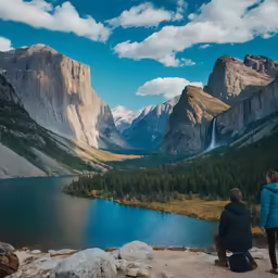 two people sit on the edge of a mountain looking at a lake and forest