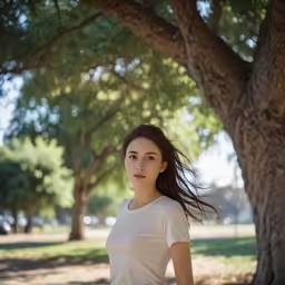 a woman in a white shirt is standing in the shade of a tree