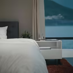 a bed with a night stand beside it in front of an ocean
