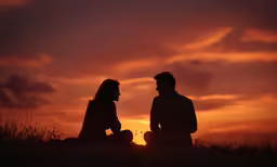 two people sitting in the grass watching a sunset