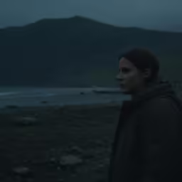 a young woman looks off to the distance while standing in a field near mountains
