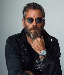 a bearded man in black jacket with his arms crossed,a man with glasses and a beard poses for a picture
