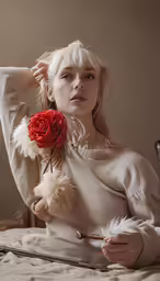 a blond woman in a white sweater is on a bed with her hand resting on a rose