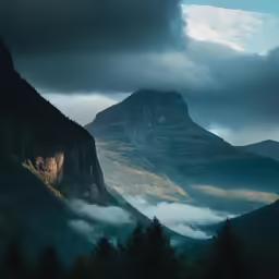 a mountain side with clouds and trees in the foreground