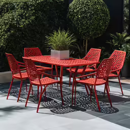 a red plastic patio table and six chairs in front of some bushes