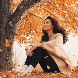 a beautiful woman sits under the autumn tree with her feet in the leaves