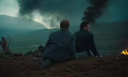 two men sitting at the edge of a field looking at an explosion