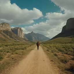 a person with backpack walking down a dirt road in front of mountains
