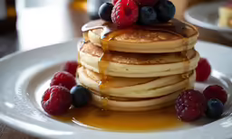 a stack of pancakes, some syrup and fruit are on a plate