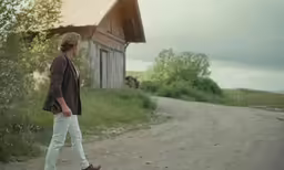 a woman wearing white jeans walking along a road next to a house