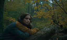 a young woman in a brown and blue jacket leans on a tree branch