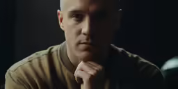 a man in a brown shirt stares into the camera