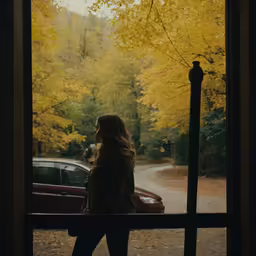 a woman stands in front of a window with autumn foliage outside