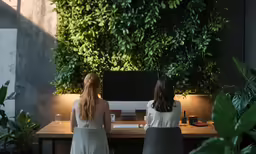two women standing at a desk in front of a plant