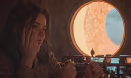 the girl holds her hand over her eyes as she sits in front of a round window