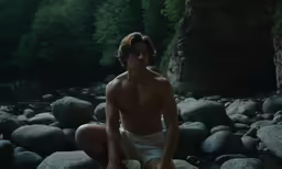 shirtless man sitting on rocks near river