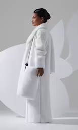 a woman in a white coat stands against a white wall
