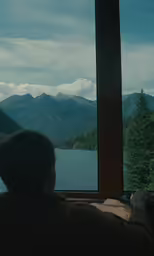 man looking out window at a lake and mountains outside