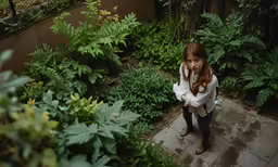 a young girl in a white jacket is standing by green plants