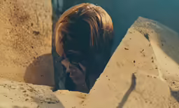 woman with red hair peering out through wall next to rocks