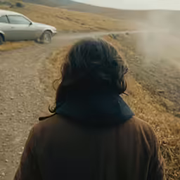 person in brown jacket walking toward a car on a dirt road