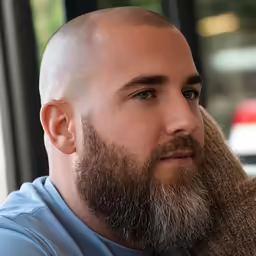 man with shaved head and beard sitting in front of large windows