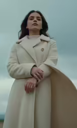 a woman stands in the middle of an open field wearing a cream coat and turtle - neck sweater