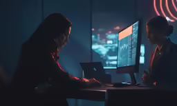 two woman in a dark office look at a computer screen