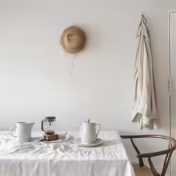 a table with a teapot, cream plate, cups, and a hat hangs above it on the wall