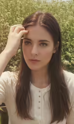 a woman in a field, with long brown hair, touching her temple
