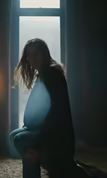a pregnant woman seated on a bed looking out an open window