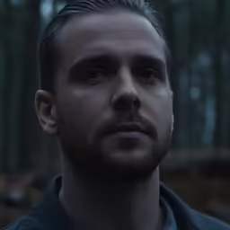a man with a goatee stares ahead in the forest